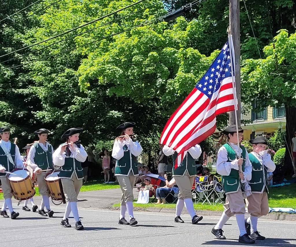 Young boys in period costumes marching in Ridgefield parade with musical instruments and United States flag.